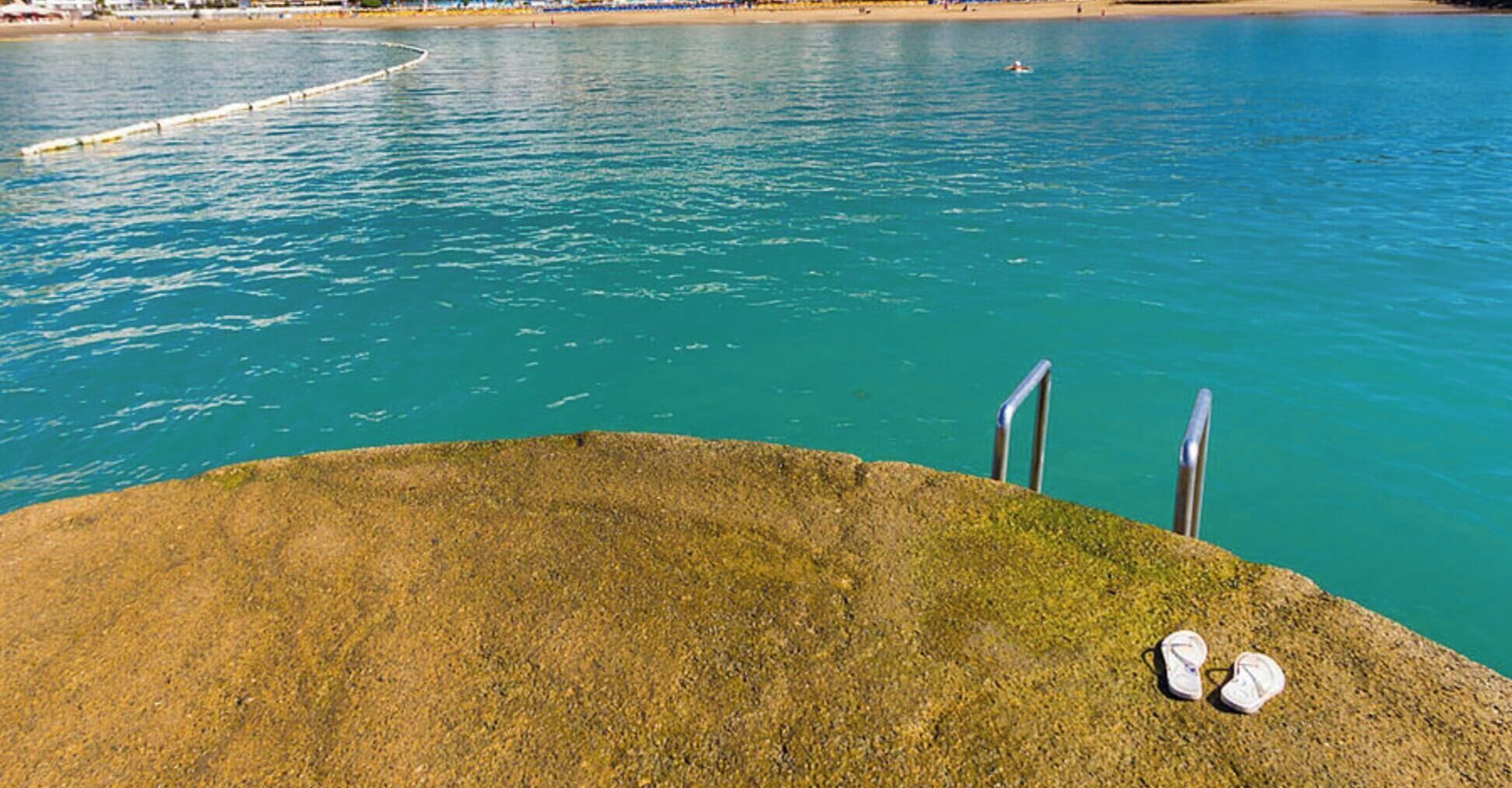 Una piscina con agua azul cristalina, fondo de arena y dos escaleras de metal.