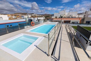 A rooftop swimming pool with clear blue water, surrounded by metal railings and a view of a town in the background.