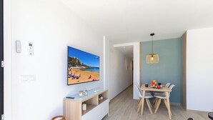 A modern living room with a TV displaying a beach scene, a dining area with a table and chairs, and a wicker lamp.