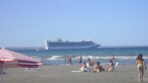 A beach with a large cruise ship in the background, people relaxing on the sand, and a red and white striped umbrella. - Hostel Viajeros (Puerto Madryn)