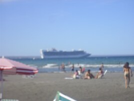 Una playa con un gran crucero al fondo, gente relajándose en la arena y un paraguas a rayas rojas y blancas.