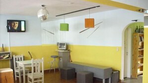 A room with a yellow and white color scheme, a television, a desk with chairs, a bench, and a shelf with bottles. - Hostel Viajeros (Puerto Madryn)