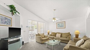 A living room with a flat-screen TV, two sofas, a glass coffee table, and a ceiling fan.