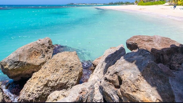A beach with clear turquoise water, large rocks in the foreground, and a small building with palm trees in the background. - Welcome to Ocean's Tropical Escape!
Beach front, beach access 1-bedroom, 4 bed. (Runaway Bay)