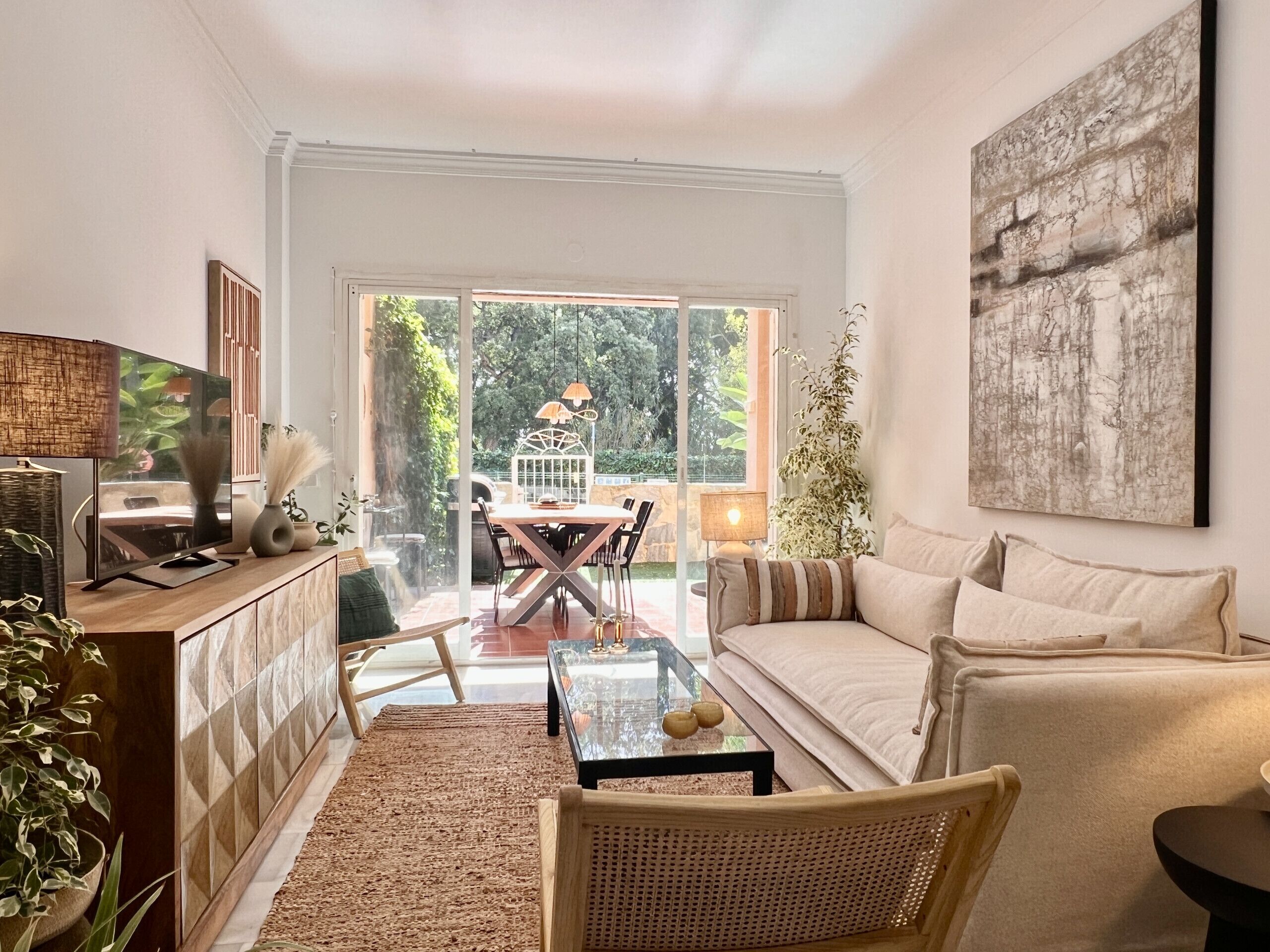 A modern living room with a sofa, a coffee table, and a TV on a cabinet. There is a dining table with chairs outside a glass door.