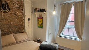 Une chambre avec un mur en briques, un lit avec une literie beige, une petite étagère avec des livres, une affiche accrochée au mur et une fenêtre avec des rideaux.