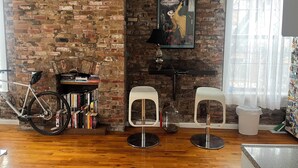 Une pièce avec un mur en briques, un vélo, une bibliothèque, deux chaises blanches, une table et une affiche.