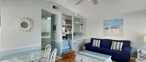 A living room with a blue sofa, white chairs, a glass table, a ceiling fan, and a painting of a beach scene.