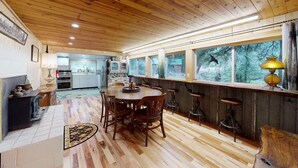 A wooden-paneled room with a dining table and chairs, a kitchen in the background, and large windows with a view of trees.