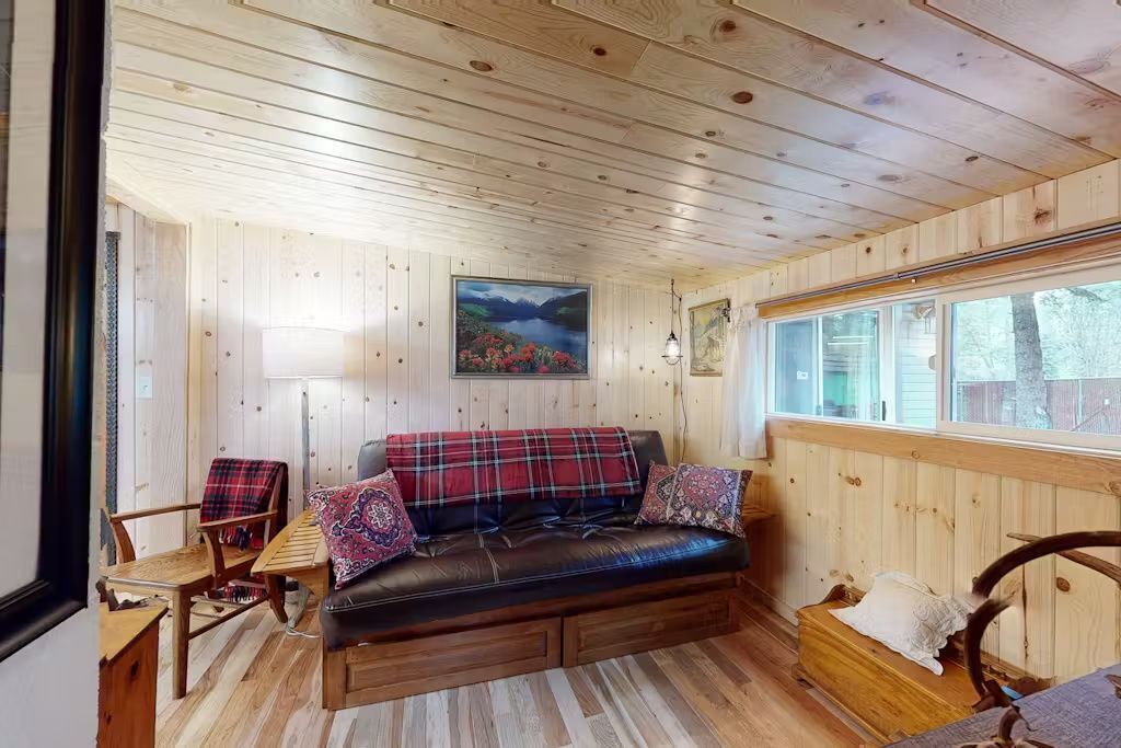 A cozy cabin interior with a wooden ceiling, a black leather sofa with plaid cushions, a wooden bench, and a painting of a landscape.