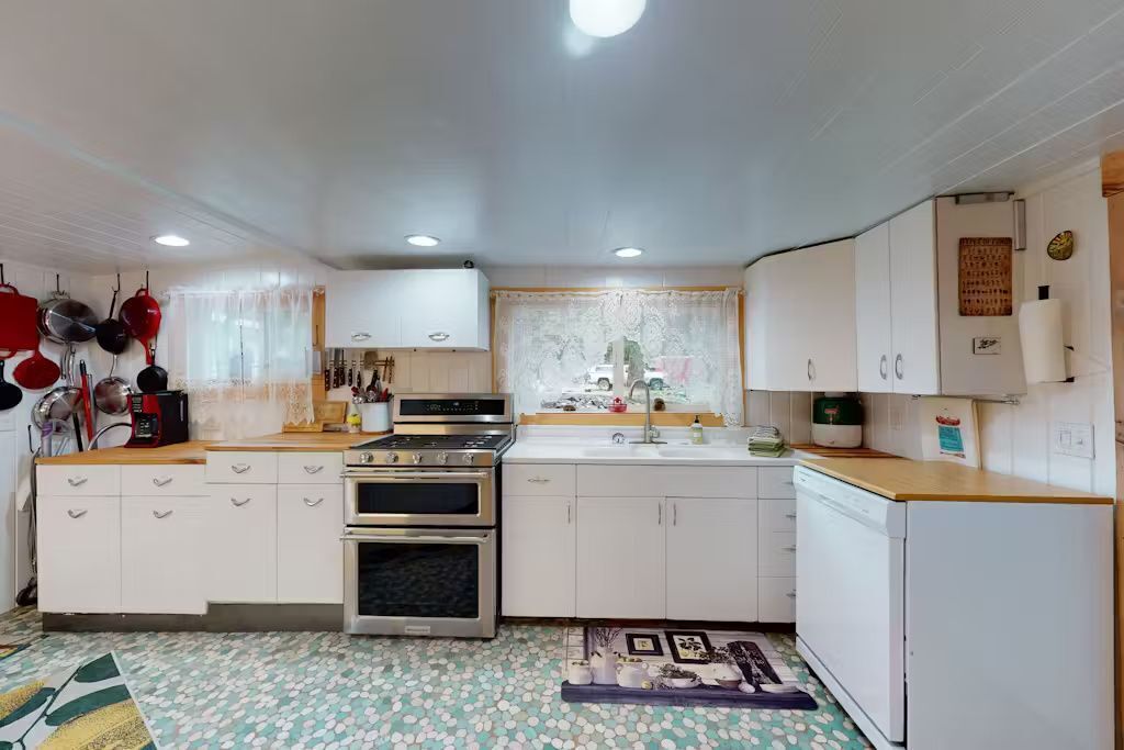 Una cucina compatta con mobili bianchi, un forno doppio, una lavastoviglie e una finestra con tende di pizzo.