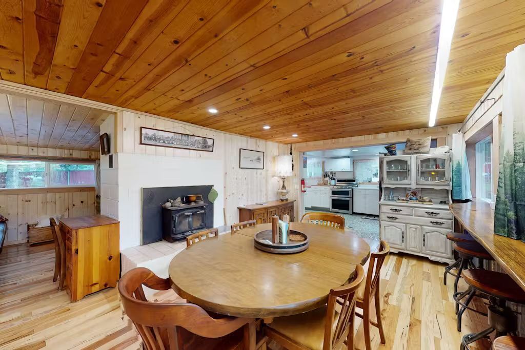 A cozy cabin interior with a wooden dining table surrounded by chairs, a fireplace, and a kitchen area visible through an open doorway.