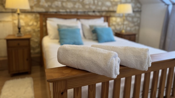 Une chambre avec un mur en pierre, une tête de lit en bois, un lit avec du linge de lit blanc, deux oreillers bleus et deux lampes de chevet.
