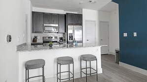 A modern kitchen with dark cabinets, stainless steel appliances, and a marble countertop. There are three bar stools and a door leading to another room. - Landing | Chic 1BD, Gym, Pool (Manor)