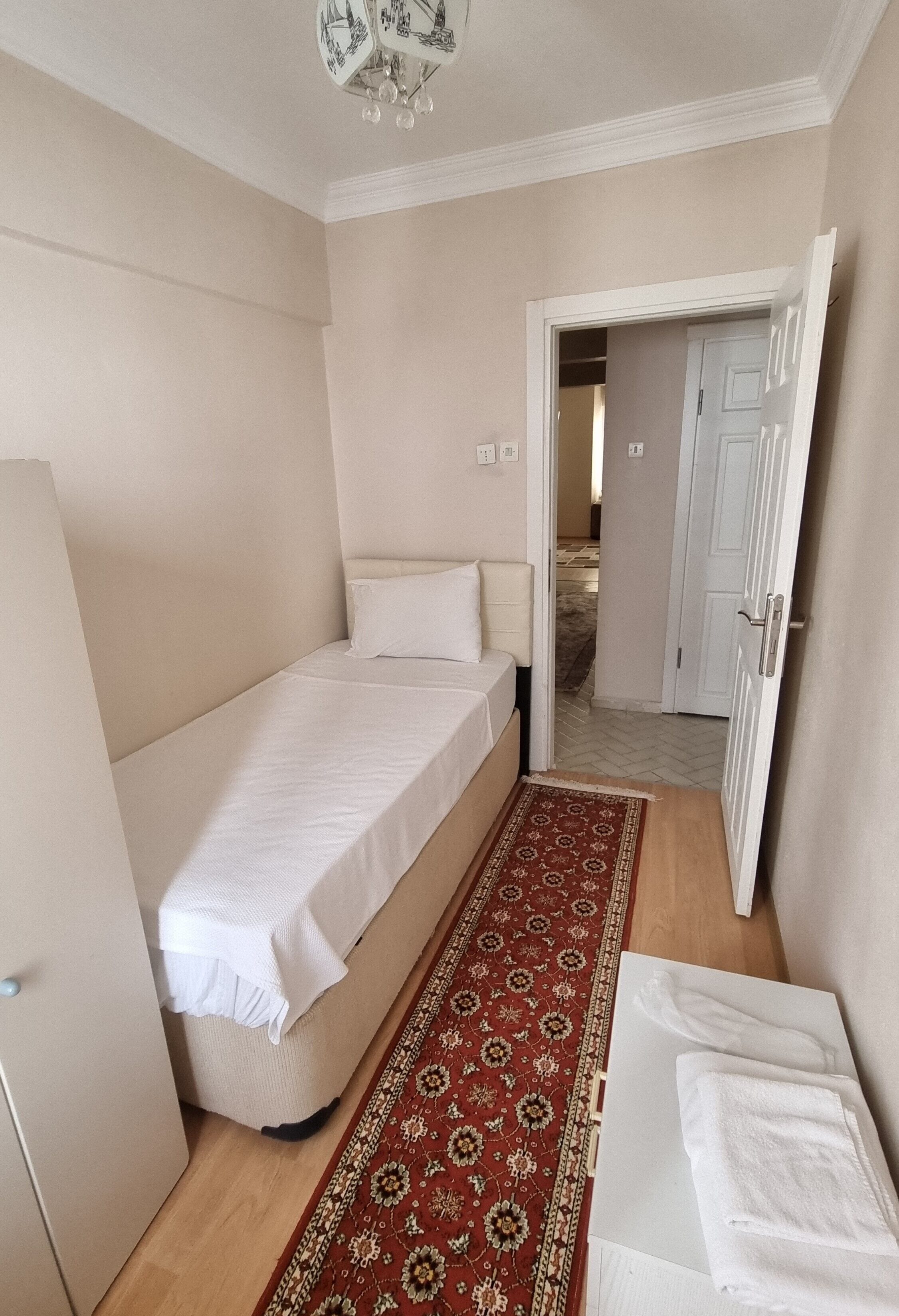 A small, neatly arranged bedroom with a single bed, a bedside table with a towel, and a patterned rug on the floor.