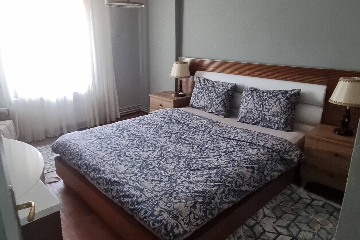 A neatly arranged hotel room with a large bed, two bedside tables with lamps, a small desk with a chair, and a patterned rug.