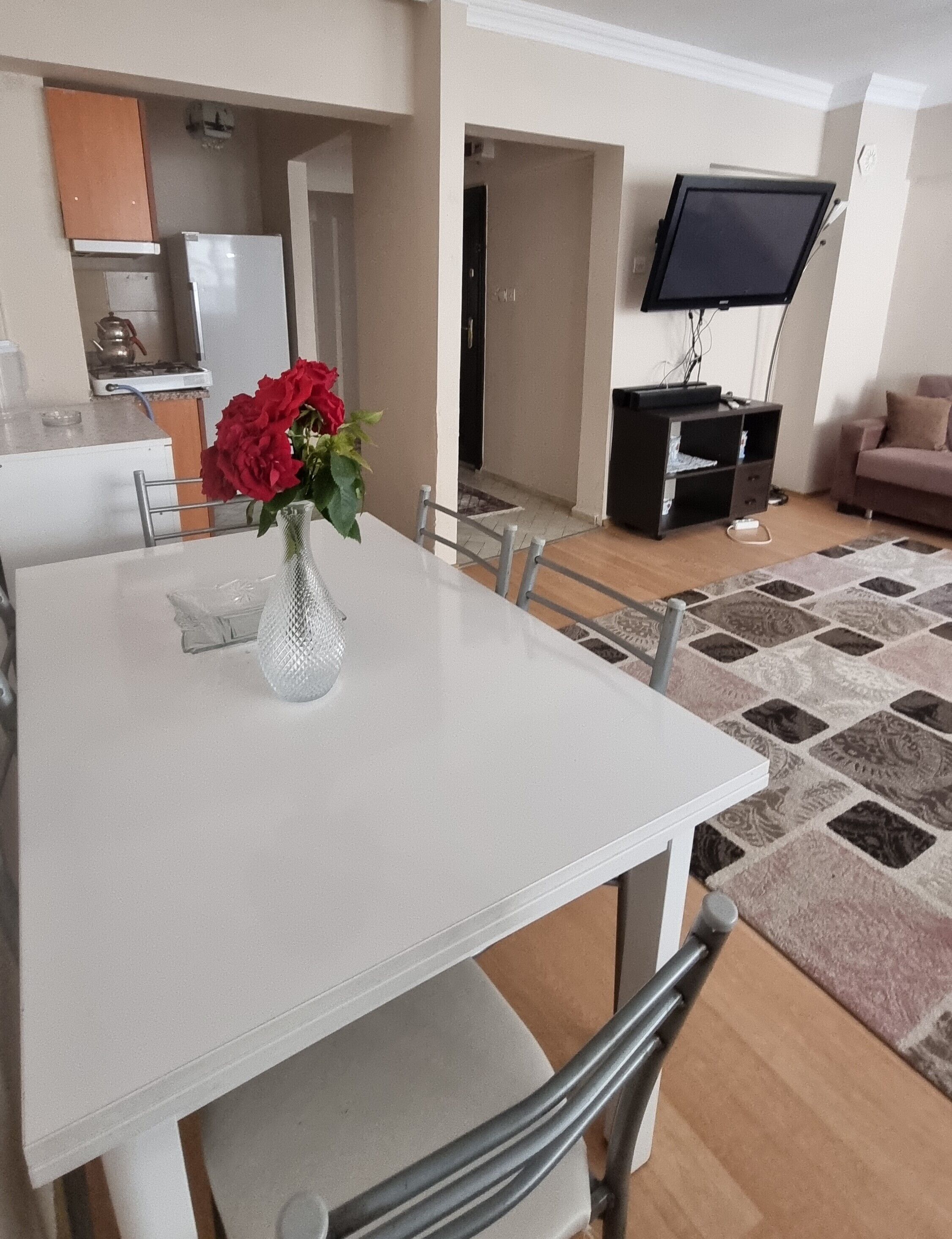 A dining area with a white table and chairs, a vase with red flowers, a kitchen in the background, a television on the wall, and a patterned rug.