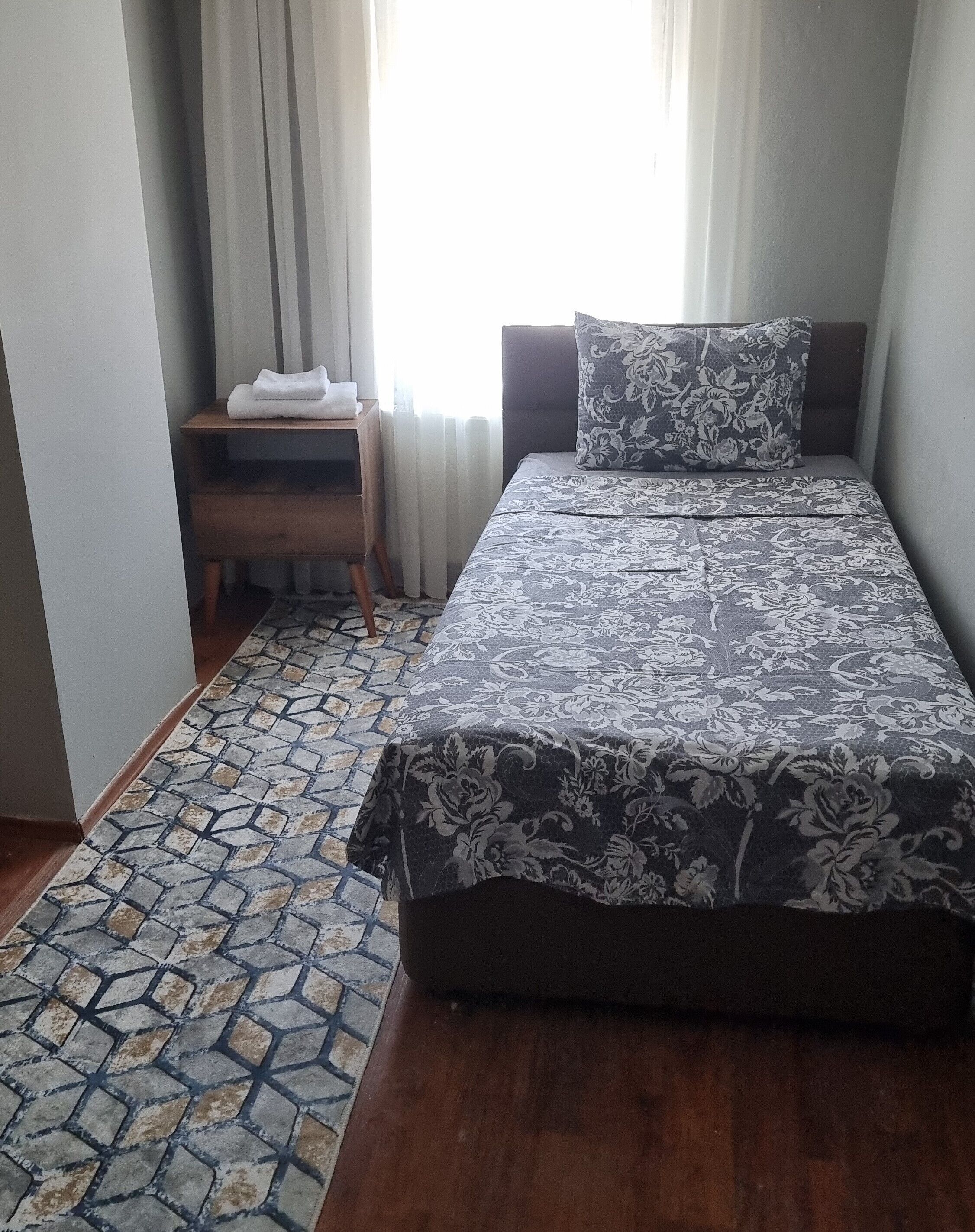 A bedroom with a bed, a nightstand, a patterned rug, and a window with curtains.