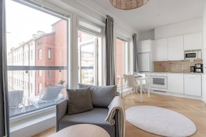 A modern apartment with a grey sofa, a dining table, a kitchen with white cabinets, and large windows offering a view of the city.
