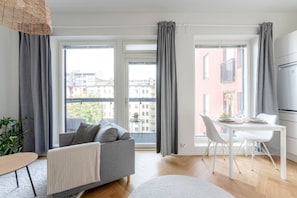 A modern apartment with a grey sofa, a dining table with chairs, and large windows with grey curtains.