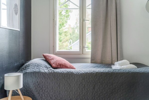 Un dormitorio con cama, ventana con cortinas, lámpara de noche y una toalla sobre la cama.