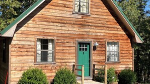 A log cabin with a green door, two windows, and a porch with potted plants.