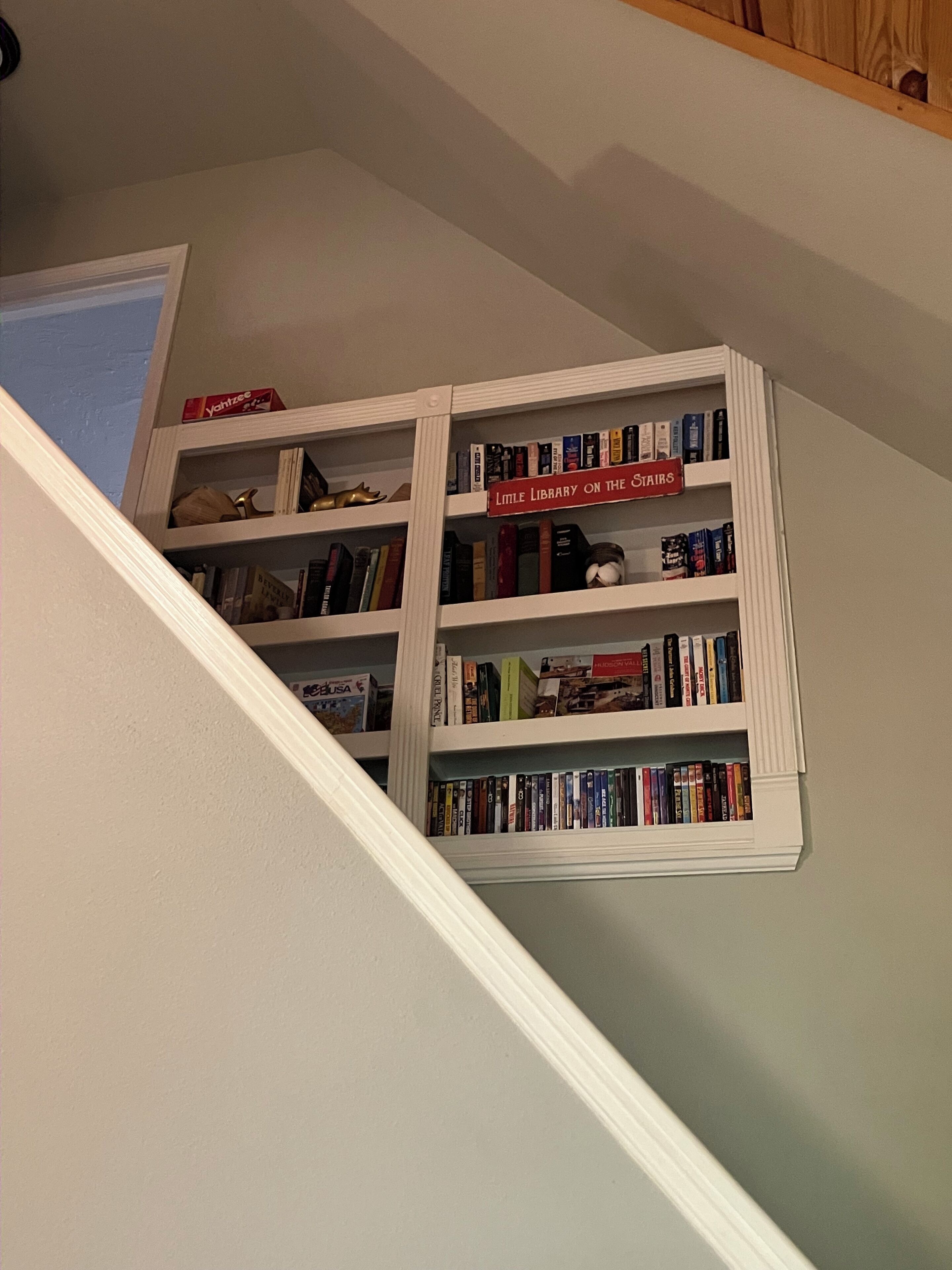 Rincón de una habitación con una escalera blanca y una estantería que dice 'Little Library on the Stairs'.