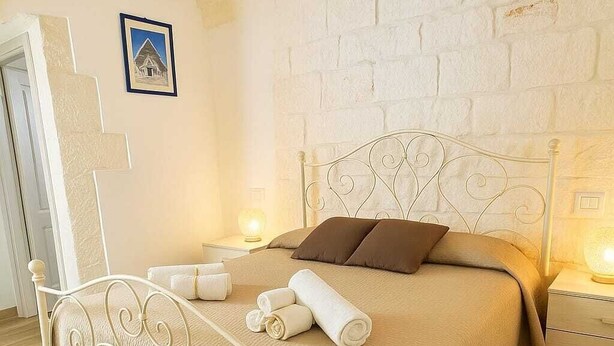 Una cama bien hecha con estructura metálica blanca, un edredón color beige y dos almohadas marrones. Hay dos mesitas de noche con lámparas, y un cuadro enmarcado en la pared.