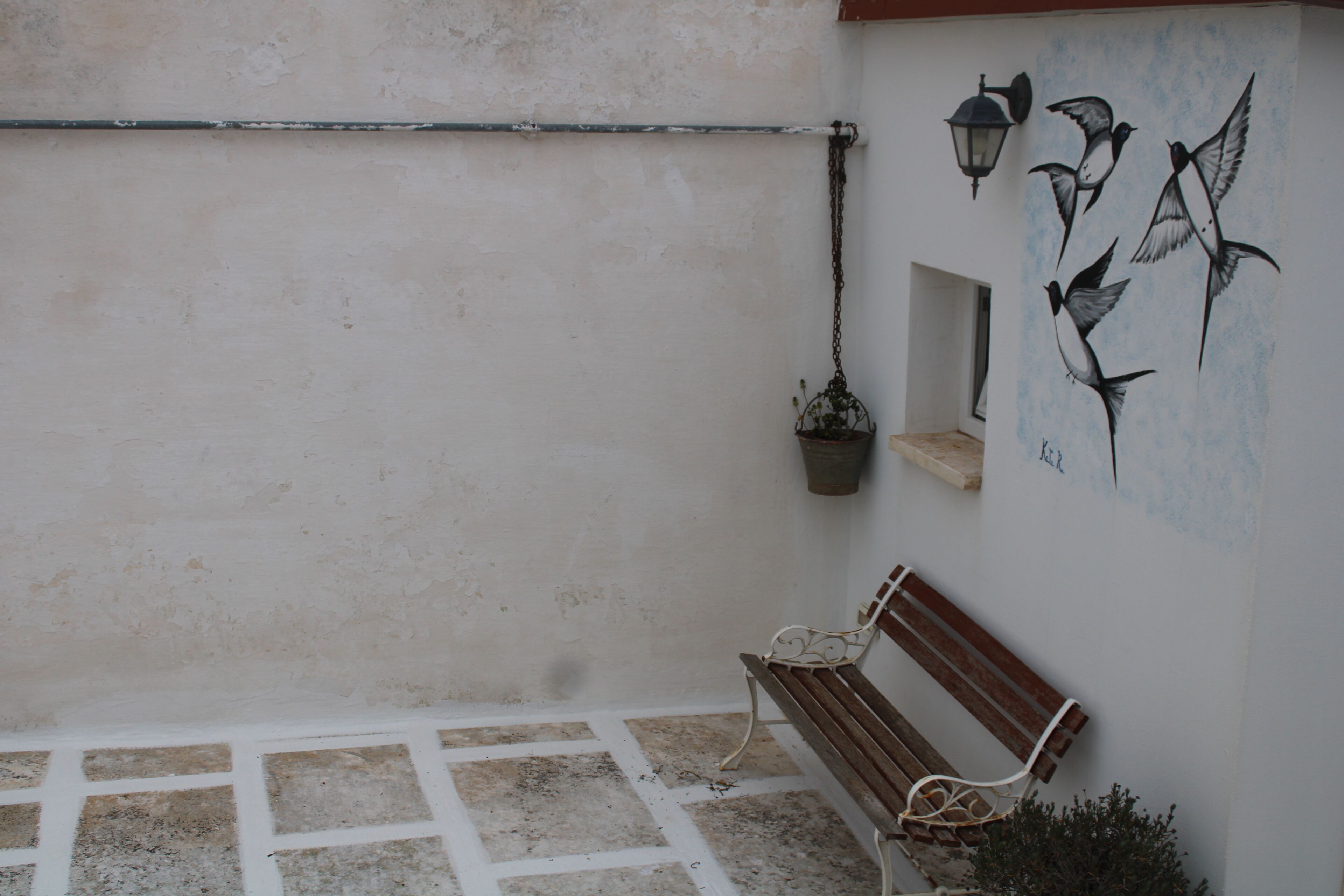 Un banc dans une cour, avec une fresque murale représentant des oiseaux, un pot de fleurs suspendu et une lanterne fixée au mur.