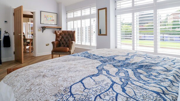 A bedroom with a large bed, a wooden chair, a mirror, and a window with blinds. - The Canalside Cottage (Derby)