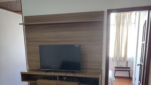 A living room with a wooden herringbone floor, a flat-screen TV mounted on a wooden cabinet, a white ottoman, and a small table with a chair. - Charming apartment in enjoyable Flamengo Rio de Janeiro near Park Aterro (Flamengo Rio de Janeiro)
