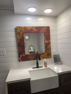 Une salle de bain avec un lavabo blanc, un robinet noir, un miroir orné d’un motif floral coloré et un petit comptoir blanc sur lequel se trouve un distributeur de savon.