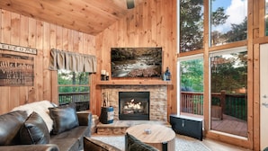 A cozy cabin interior with a fireplace, leather sofa, and a large window overlooking a deck. - Mountainside retreat in the Smokies!  6 miles to The Island. Sleeps 8. (Sevierville)