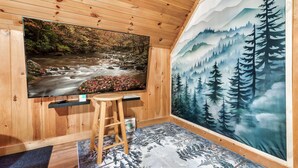 A room with a large TV on a wooden wall, a wooden stool, and a mural of a forest and mountains. - Mountainside retreat in the Smokies!  6 miles to The Island. Sleeps 8. (Sevierville)
