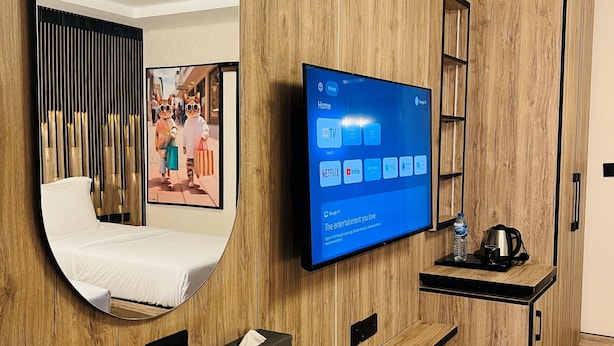 A hotel room with a wooden wall, a large mirror, a flat-screen TV displaying a home screen, a bed, a desk with a chair, and a wardrobe. - The Westbury Inn Hotels and Apartments (Abuja)