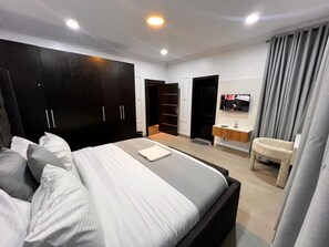 Un dormitorio moderno con una cama grande, un armario oscuro, una tocador con espejo y un televisor.