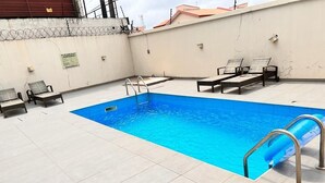Ein rechteckiger Pool mit einer Metallleiter an einer Seite, umgeben von hell gefliestem Boden. Am Pool befinden sich zwei Liegestühle und ein kleiner Tisch.