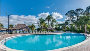 A clear blue pool surrounded by lounge chairs and palm trees, with a thatched-roof structure in the background.
