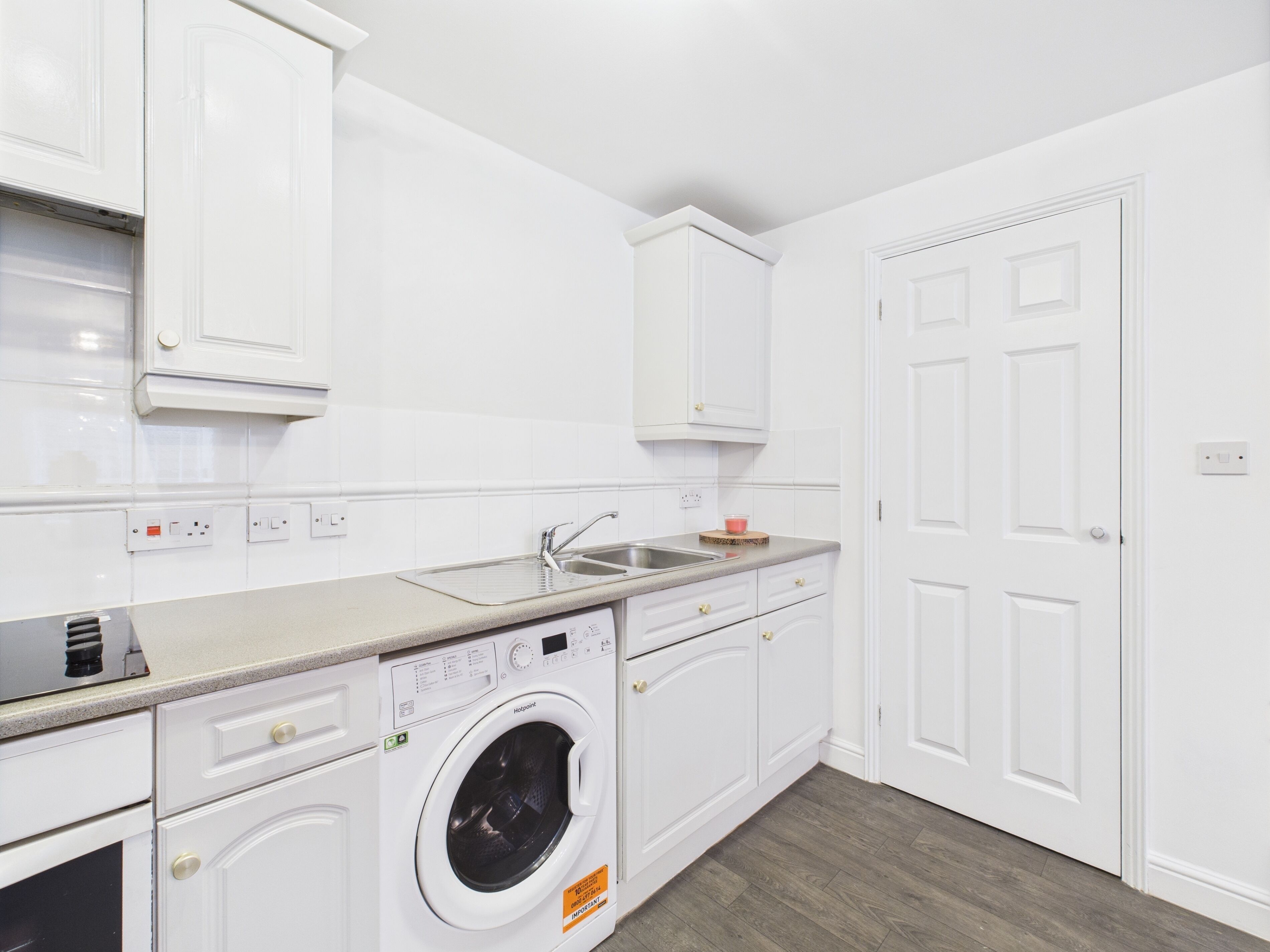 A compact kitchen with white cabinetry, a built-in washing machine, a countertop with a sink, and a wooden floor.