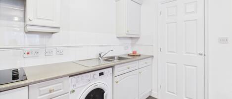 A compact kitchen with white cabinetry, a built-in washing machine, a countertop with a sink, and a wooden floor.