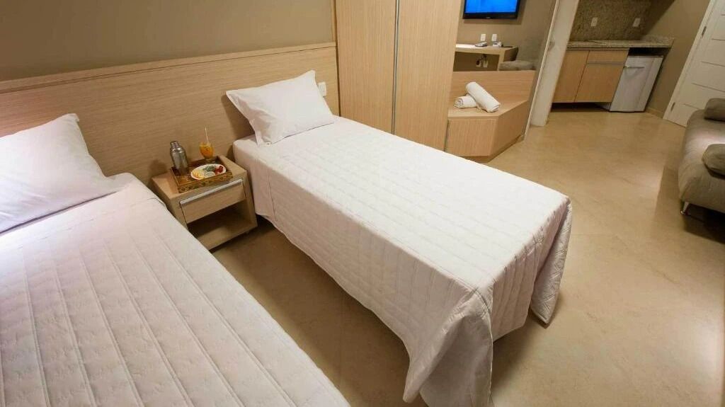 A hotel room with two single beds, a small bedside table with a bottle and a glass, a TV mounted on the wall, and a kitchenette area with a microwave and a small fridge.