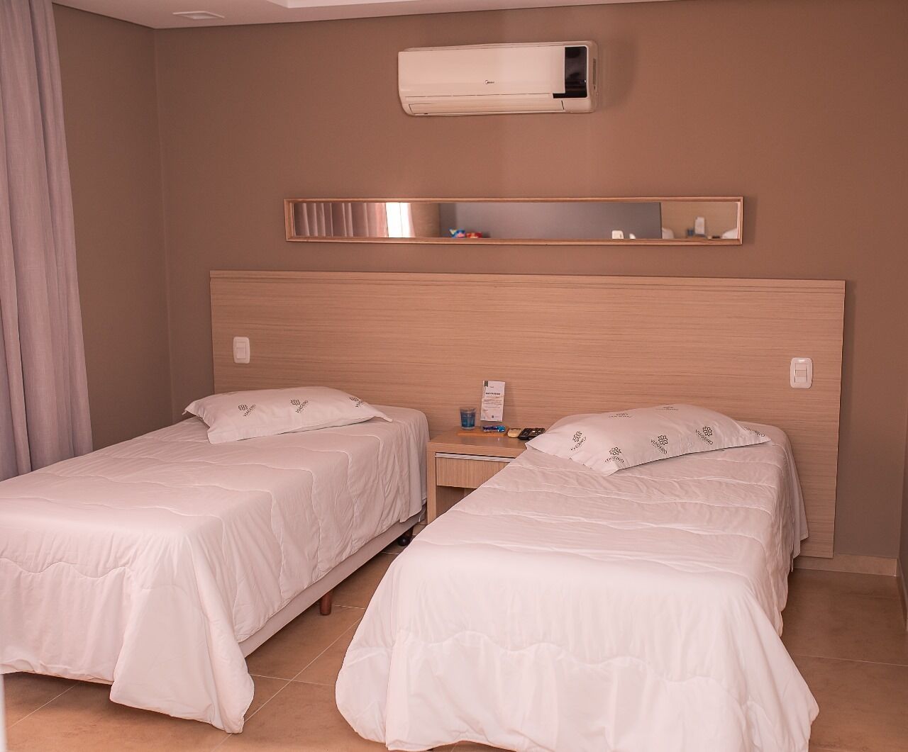 Habitación de hotel con dos camas, una mesita de noche, un equipo de aire acondicionado y un espejo sobre la cabecera.