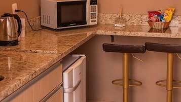 A compact kitchen with a microwave, a kettle, a coffee maker, a sink, and a trash can. There are two stools and a basket of snacks on the counter.