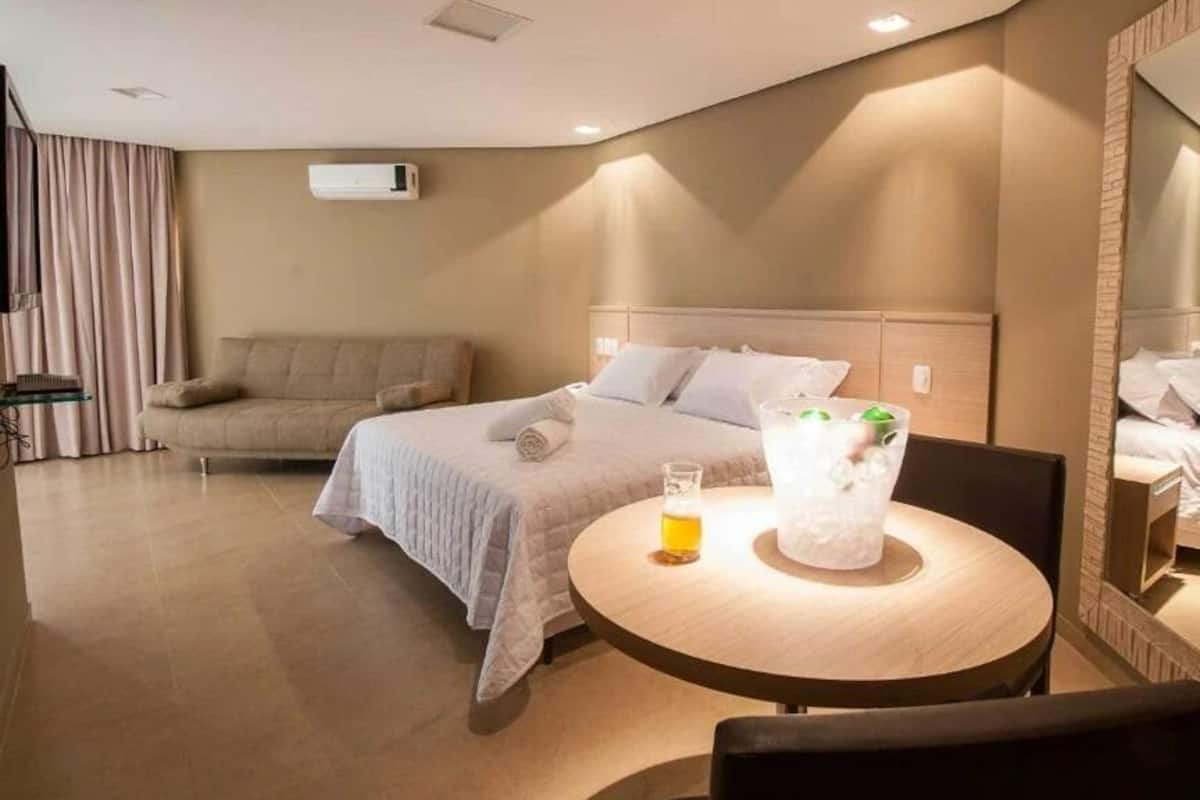 A hotel room with a bed, a round table with a glass of beer, a sofa, and a large mirror.