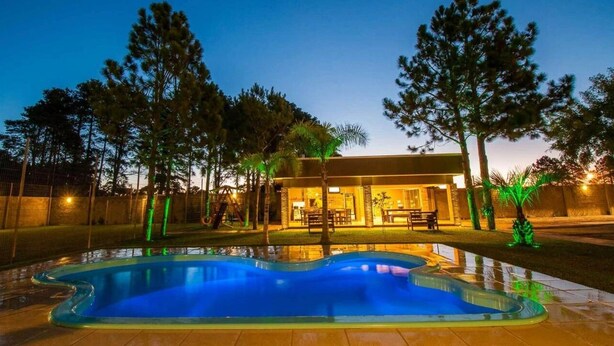 A swimming pool with a wavy edge, surrounded by trees and a building with a patio area. - Viageiro Hotel (Pelotas)