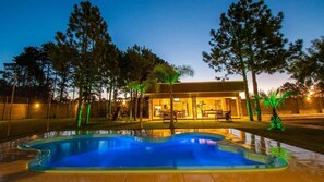 A swimming pool with a wavy edge, surrounded by trees and a building with a patio area. - Viageiro Hotel (Pelotas)
