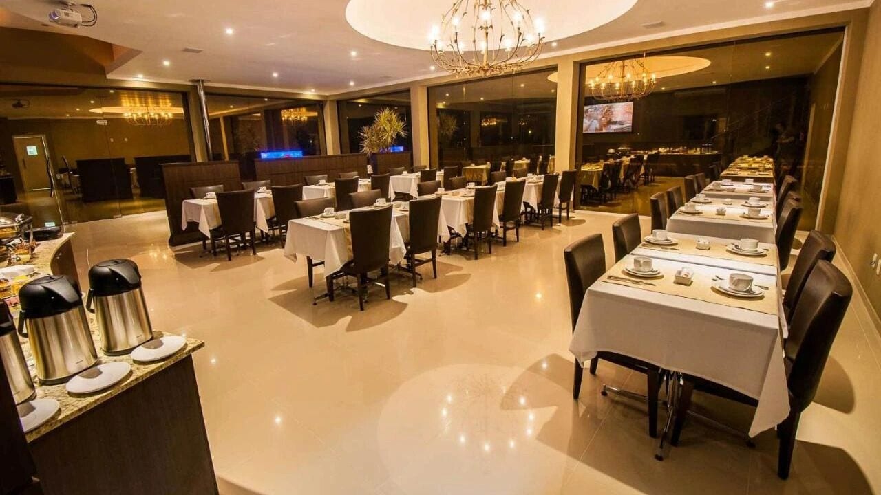 A spacious dining area with multiple tables and chairs, a chandelier, and a television screen in the background.