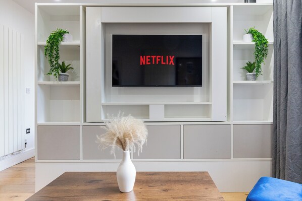 A modern living room with a flat-screen TV mounted on a white wall, surrounded by shelves with plants.