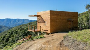 A wooden cabin with a deck, situated on a dirt path with a mountainous backdrop. - Cabana c\Jacuzzi prox. de Florianópolis VST014 (Anitapolis)