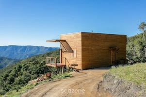 Una cabaña de madera con un balcón, ubicada en un sendero de tierra con montañas al fondo.